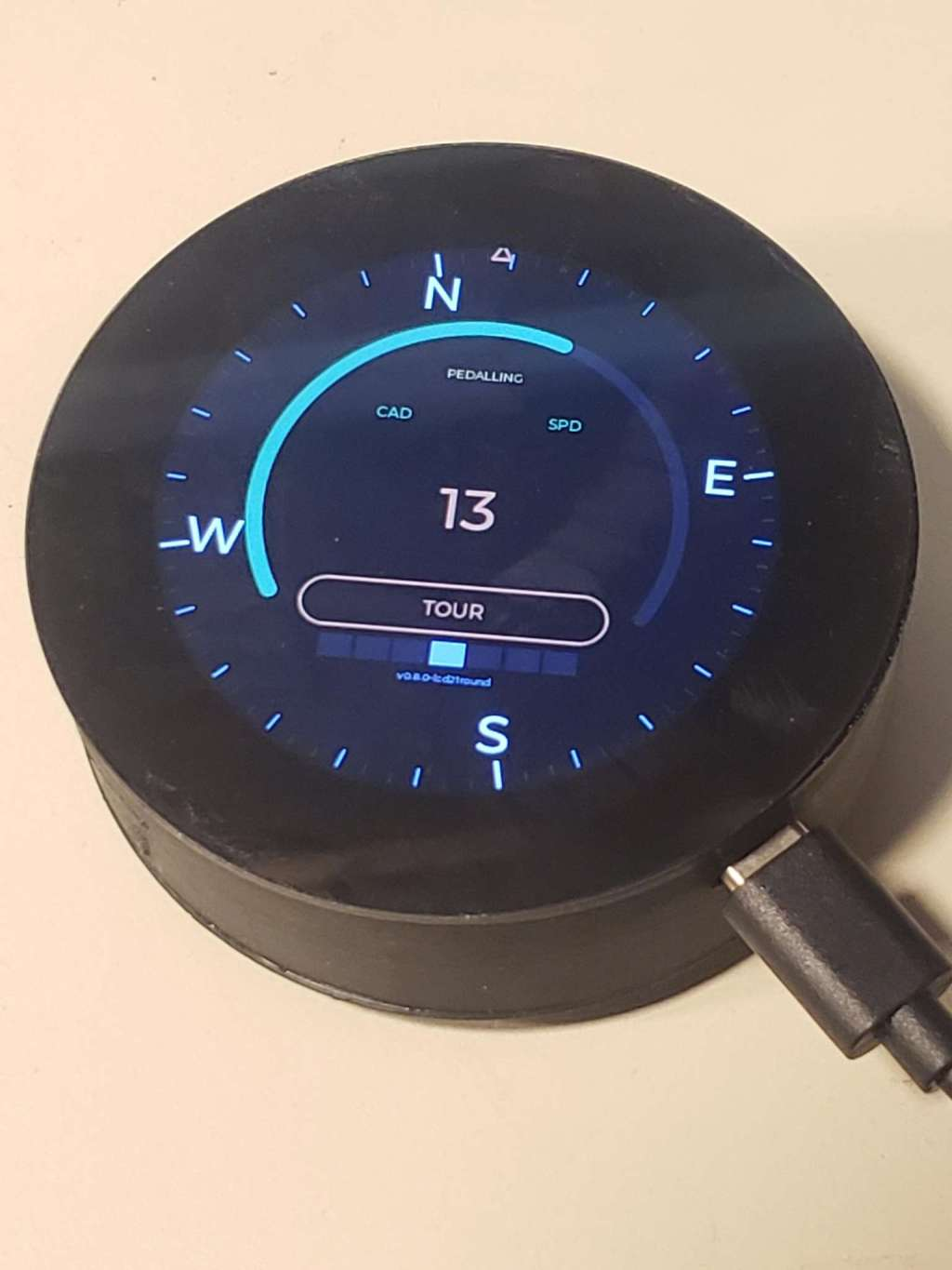 GearSense TIM-less round display unit showing circular UI with compass bezel, cyan effort arc, speed reading of 13, TOUR mode, and PEDALLING status. Version v0.8.0-lcd21round.